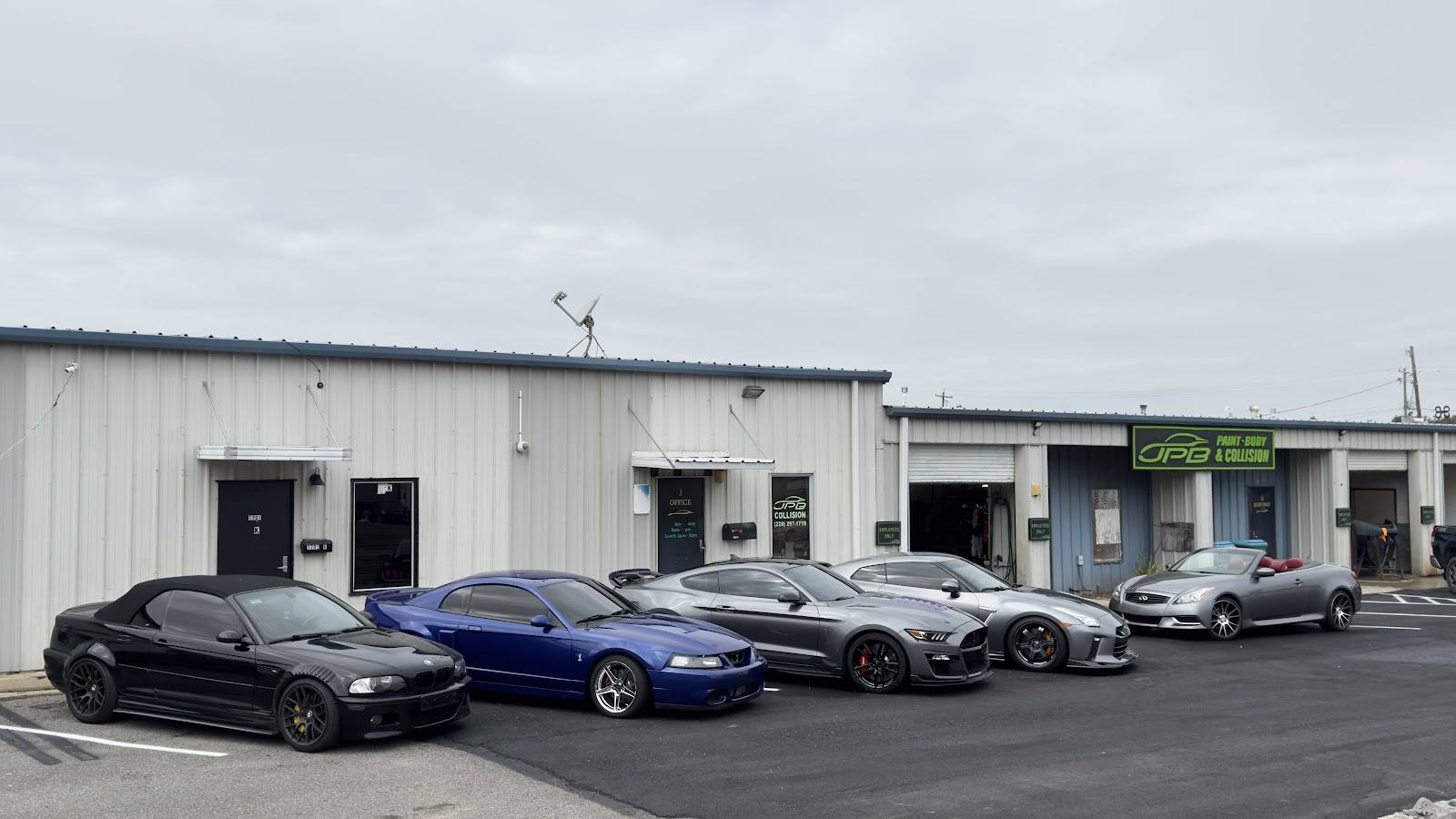 Five sports cars parked in a row outside a metal shop building with a green JPB Paint Body & Collision sign